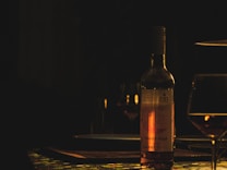 A dimly lit scene featuring a bottle of Merlot Ros&eacute; wine on a tablecloth with geometric patterns. The bottle's label is partially illuminated, casting a warm glow. There are glasses partially filled with wine set around, creating an intimate and cozy ambiance.