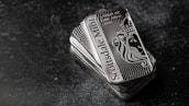 A stack of shiny rectangular silver bars with intricate designs and the inscription 'Scottsdale Mint'. The bars feature a depiction of a lion and have text indicating purity and weight, 'FINE Silver .999'. The background is a dark textured surface, providing contrast to the reflective nature of the bars.