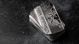 A stack of shiny rectangular silver bars with intricate designs and the inscription 'Scottsdale Mint'. The bars feature a depiction of a lion and have text indicating purity and weight, 'FINE Silver .999'. The background is a dark textured surface, providing contrast to the reflective nature of the bars.