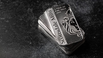 A stack of shiny rectangular silver bars with intricate designs and the inscription 'Scottsdale Mint'. The bars feature a depiction of a lion and have text indicating purity and weight, 'FINE Silver .999'. The background is a dark textured surface, providing contrast to the reflective nature of the bars.