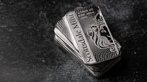 A stack of shiny rectangular silver bars with intricate designs and the inscription 'Scottsdale Mint'. The bars feature a depiction of a lion and have text indicating purity and weight, 'FINE Silver .999'. The background is a dark textured surface, providing contrast to the reflective nature of the bars.