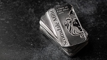 A stack of shiny rectangular silver bars with intricate designs and the inscription 'Scottsdale Mint'. The bars feature a depiction of a lion and have text indicating purity and weight, 'FINE Silver .999'. The background is a dark textured surface, providing contrast to the reflective nature of the bars.