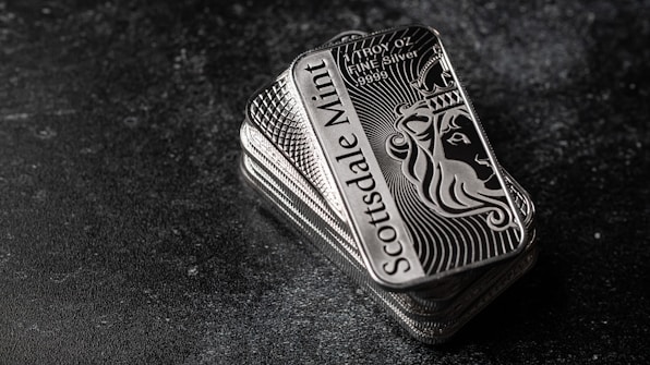 A stack of shiny rectangular silver bars with intricate designs and the inscription 'Scottsdale Mint'. The bars feature a depiction of a lion and have text indicating purity and weight, 'FINE Silver .999'. The background is a dark textured surface, providing contrast to the reflective nature of the bars.