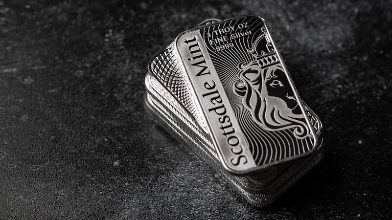 A stack of shiny rectangular silver bars with intricate designs and the inscription 'Scottsdale Mint'. The bars feature a depiction of a lion and have text indicating purity and weight, 'FINE Silver .999'. The background is a dark textured surface, providing contrast to the reflective nature of the bars.