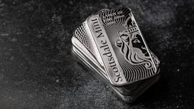 A stack of shiny rectangular silver bars with intricate designs and the inscription 'Scottsdale Mint'. The bars feature a depiction of a lion and have text indicating purity and weight, 'FINE Silver .999'. The background is a dark textured surface, providing contrast to the reflective nature of the bars.