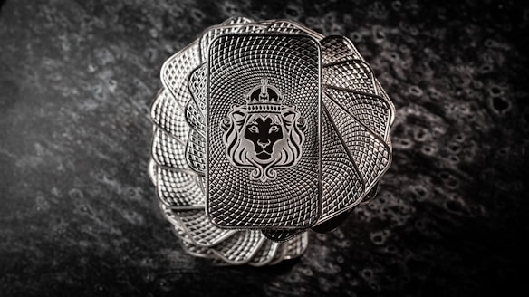 A stack of intricately designed rectangular silver bars with a lion's head wearing a crown engraved in the center. The bars have a textured pattern resembling a spiral, creating an elegant and regal appearance. The background is a dark, marbled surface that enhances the metallic shine of the bars.