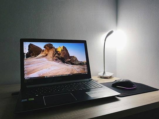 A laptop on a wooden desk displays a scenic rock formation wallpaper. A sleek desk lamp illuminates the area from the right, casting soft light on the wall. A black gaming mouse sits on a mouse pad beside the laptop, creating a modern and minimalist workspace.