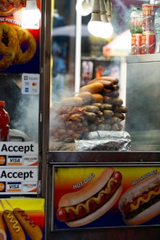 A street food cart is displaying a variety of hot dogs and sausages. There is visible steam indicating freshly cooked food. The cart is adorned with colorful images of pretzels and hot dogs, suggesting menu options. Payment signs for credit card acceptance are visible. Bright lighting comes from overhead bulbs, and some canned food items are stored on top of the cart.