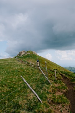 A hiker is walking along a narrow path flanked by a rustic, old wooden fence. The path winds up a grassy hillside towards a rocky peak under a sky filled with dramatic, dark clouds, while patches of sunlight illuminate the vibrant green landscape.