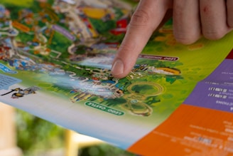 A hand with a finger pointing at a colorful map, which appears to be a theme park or amusement park layout. The map is detailed with various attractions, pathways, and labeled areas, surrounded by vibrant colors and illustrations.