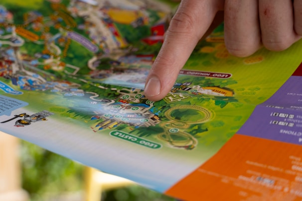 A hand with a finger pointing at a colorful map, which appears to be a theme park or amusement park layout. The map is detailed with various attractions, pathways, and labeled areas, surrounded by vibrant colors and illustrations.