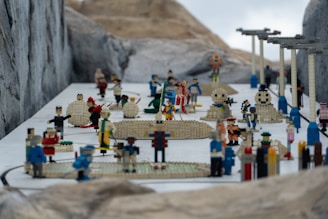 A detailed scene featuring numerous Lego figures arranged in various poses, illustrating a miniature snow-covered landscape. The figures are engaging in a range of activities, with some involved in snowball fights and others building snowmen. Architectural structures, made from Lego bricks, are scattered throughout the scene, adding to the complexity. The environment suggests a playful winter theme.