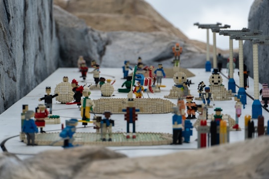 A detailed scene featuring numerous Lego figures arranged in various poses, illustrating a miniature snow-covered landscape. The figures are engaging in a range of activities, with some involved in snowball fights and others building snowmen. Architectural structures, made from Lego bricks, are scattered throughout the scene, adding to the complexity. The environment suggests a playful winter theme.