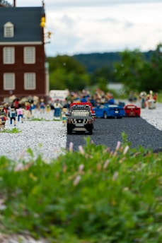 A miniature scene featuring LEGO figures and vehicles in a detailed diorama. The foreground has a road with a LEGO car driving toward the viewer, and people are gathered behind the vehicle. A LEGO building is visible in the background against a partly cloudy sky, and lush greenery frames the bottom of the image.