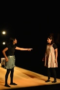 Two individuals are engaged in an intense interaction on a stage. One person, wearing a black top and patterned skirt, is pointing emphatically at the other, who is dressed in a striped dress with dark leggings. They are illuminated by stage lights against a dark background, suggesting a theatrical performance.