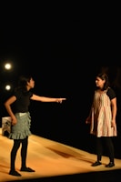 Two individuals are engaged in an intense interaction on a stage. One person, wearing a black top and patterned skirt, is pointing emphatically at the other, who is dressed in a striped dress with dark leggings. They are illuminated by stage lights against a dark background, suggesting a theatrical performance.