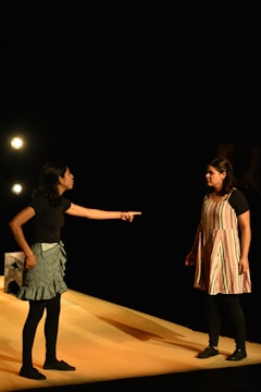 Two individuals are engaged in an intense interaction on a stage. One person, wearing a black top and patterned skirt, is pointing emphatically at the other, who is dressed in a striped dress with dark leggings. They are illuminated by stage lights against a dark background, suggesting a theatrical performance.