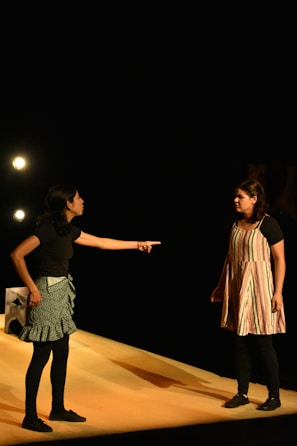 Two individuals are engaged in an intense interaction on a stage. One person, wearing a black top and patterned skirt, is pointing emphatically at the other, who is dressed in a striped dress with dark leggings. They are illuminated by stage lights against a dark background, suggesting a theatrical performance.