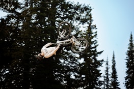A person is performing an impressive upside-down stunt on a mountain bike surrounded by tall pine trees. The rider, dressed in a white jersey and protective gear, is airborne in a forest setting with a clear blue sky.