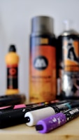A collection of art supplies including spray paint cans and markers are arranged on a surface. The focus is on the markers in the foreground, with vibrant caps in black, white, and purple. In the background, there are blurred spray cans with orange and black labels.