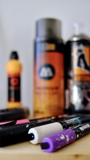 A collection of art supplies including spray paint cans and markers are arranged on a surface. The focus is on the markers in the foreground, with vibrant caps in black, white, and purple. In the background, there are blurred spray cans with orange and black labels.