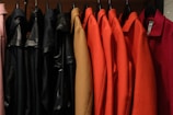 A row of coats hanging on hangers, featuring a variety of colors such as pink, black, beige, orange, and red. The coats are organized neatly in a line, and the material appears to range from fabric to leather.