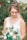 A bride dressed in a white lace gown holding a bouquet with white, green, and gold flowers. Her hair is styled in an elegant updo adorned with small floral embellishments. The background is a soft blur of green foliage and colorful flowers, creating a serene garden setting.