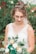 A bride dressed in a white lace gown holding a bouquet with white, green, and gold flowers. Her hair is styled in an elegant updo adorned with small floral embellishments. The background is a soft blur of green foliage and colorful flowers, creating a serene garden setting.