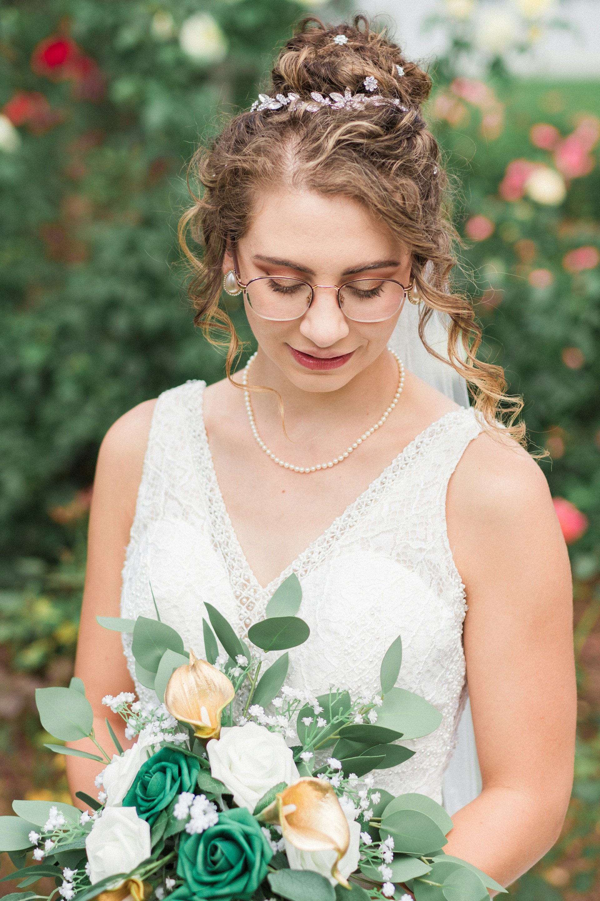 A bride dressed in a white lace gown holding a bouquet with white, green, and gold flowers. Her hair is styled in an elegant updo adorned with small floral embellishments. The background is a soft blur of green foliage and colorful flowers, creating a serene garden setting.