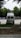 A white van is parked in an open area surrounded by trees. The vehicle is facing away, with a ladder on its rear door, and the brand 'Westfalia' is visible. To the left, several portable toilets are lined up in blue and there are cars parked in the background.