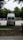 A white van is parked in an open area surrounded by trees. The vehicle is facing away, with a ladder on its rear door, and the brand 'Westfalia' is visible. To the left, several portable toilets are lined up in blue and there are cars parked in the background.
