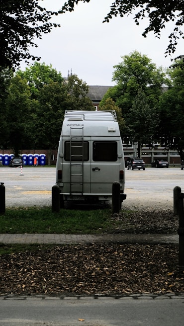 A white van is parked in an open area surrounded by trees. The vehicle is facing away, with a ladder on its rear door, and the brand 'Westfalia' is visible. To the left, several portable toilets are lined up in blue and there are cars parked in the background.