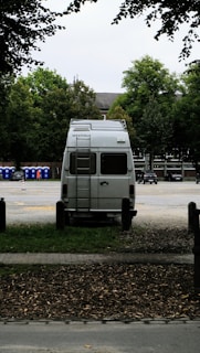 A white van is parked in an open area surrounded by trees. The vehicle is facing away, with a ladder on its rear door, and the brand 'Westfalia' is visible. To the left, several portable toilets are lined up in blue and there are cars parked in the background.