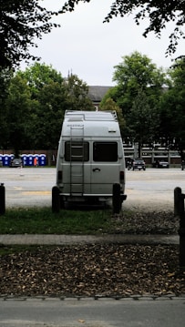 A white van is parked in an open area surrounded by trees. The vehicle is facing away, with a ladder on its rear door, and the brand 'Westfalia' is visible. To the left, several portable toilets are lined up in blue and there are cars parked in the background.