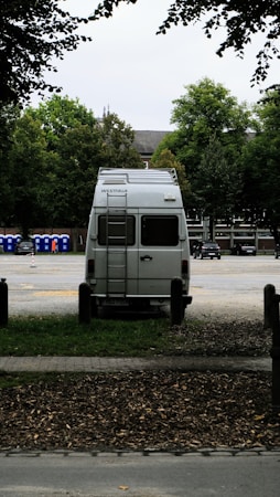 A white van is parked in an open area surrounded by trees. The vehicle is facing away, with a ladder on its rear door, and the brand 'Westfalia' is visible. To the left, several portable toilets are lined up in blue and there are cars parked in the background.