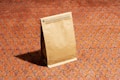 A brown paper bag stands upright on an intricately patterned orange carpet. The bag has a sealed top with a small, easily tearable opening strip. The carpet features a repetitive design in darker shades with hints of blue and purple.