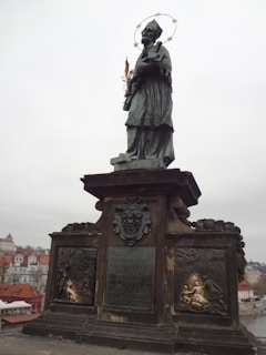 A bronze statue of a religious figure is mounted on a large, ornate pedestal. The figure appears to be a saint, wearing a robe, with a halo of stars around the head. The pedestal is adorned with detailed carvings and inscriptions, with a sculpted coat of arms in the center. In the background, there are historic buildings with red-tiled roofs, under a cloudy sky.