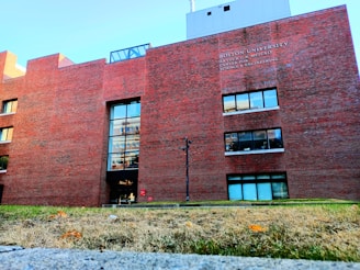 A large red brick building with several windows and a main entrance. The building's facade includes the name 'Boston University' and a description above one set of windows. The foreground features a grass area with some fallen leaves, and a clear blue sky is visible above the structure.