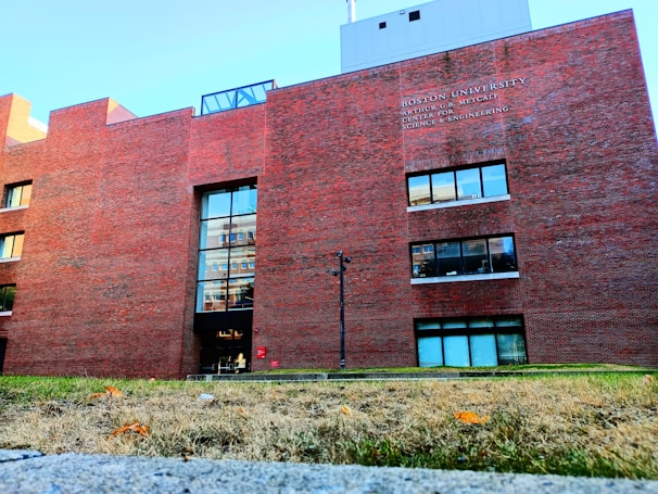 A large red brick building with several windows and a main entrance. The building's facade includes the name 'Boston University' and a description above one set of windows. The foreground features a grass area with some fallen leaves, and a clear blue sky is visible above the structure.