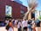 A group of people gather in an outdoor courtyard area near a modern brick building with large windows. A striking metal sculpture with long, thin rods extends upwards near the building's entrance. The people appear to be engaged in conversation or waiting for an event.