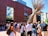 A group of people gather in an outdoor courtyard area near a modern brick building with large windows. A striking metal sculpture with long, thin rods extends upwards near the building's entrance. The people appear to be engaged in conversation or waiting for an event.