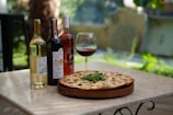 A table set outdoors showcasing a pizza with greens, olives, and cheese on a wooden board. Beside the pizza are three bottles of wine—white, red, and rosé—and a glass filled with red wine. The background is blurred with hints of greenery, suggesting a tranquil garden setting.