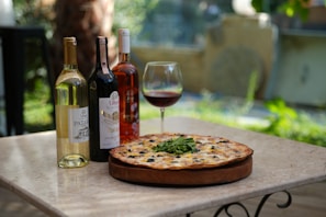 A table set outdoors showcasing a pizza with greens, olives, and cheese on a wooden board. Beside the pizza are three bottles of wine—white, red, and rosé—and a glass filled with red wine. The background is blurred with hints of greenery, suggesting a tranquil garden setting.