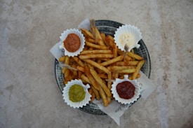 A plate filled with seasoned French fries is arranged with four small containers of dipping sauces on a textured, light-colored surface. The sauces appear to be ketchup, mayonnaise, guacamole, and a brown sauce, possibly barbecue.