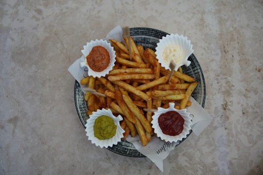 A plate filled with seasoned French fries is arranged with four small containers of dipping sauces on a textured, light-colored surface. The sauces appear to be ketchup, mayonnaise, guacamole, and a brown sauce, possibly barbecue.