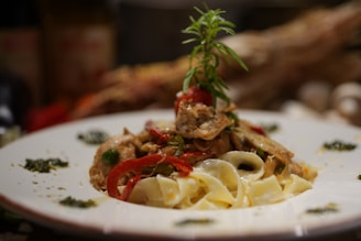 A plate of pasta topped with a mix of vegetables and herbs. The dish features red bell peppers, mushrooms, and a sprig of rosemary garnishing the top. The pasta appears creamy, and the plate is artistically decorated with small amounts of green sauce around the edges.