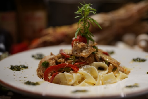 A plate of pasta topped with a mix of vegetables and herbs. The dish features red bell peppers, mushrooms, and a sprig of rosemary garnishing the top. The pasta appears creamy, and the plate is artistically decorated with small amounts of green sauce around the edges.