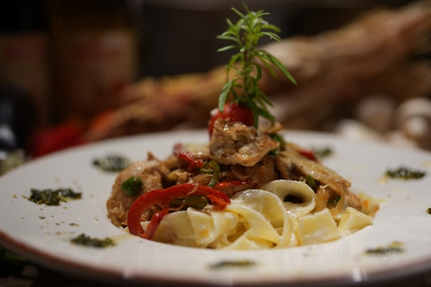 A plate of pasta topped with a mix of vegetables and herbs. The dish features red bell peppers, mushrooms, and a sprig of rosemary garnishing the top. The pasta appears creamy, and the plate is artistically decorated with small amounts of green sauce around the edges.
