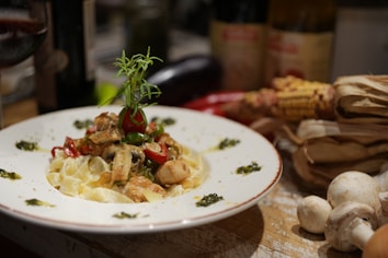 A plate of pasta topped with pieces of chicken, vegetables, and herbs garnished with a sprig of rosemary. The dish is surrounded by ingredients like mushrooms and corn, with bottles and a glass of red wine in the background.