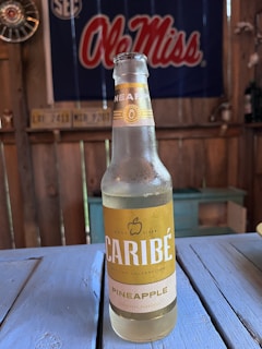 A bottle of Caribe pineapple-flavored cider sits on a blue wooden table. The bottle is clear with yellow and white labeling, featuring an apple icon. In the background, a red and white Ole Miss banner hangs on a rustic wooden wall alongside old license plates and hubcaps.
